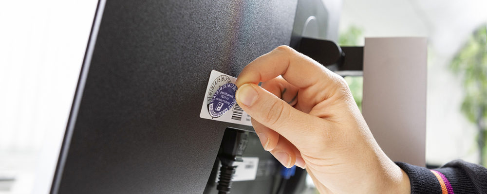 Aufkleben einer DGUV-Prüfplakette auf einen Bildschirm