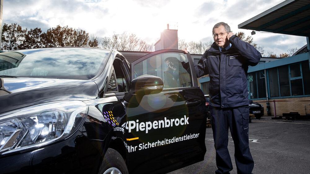 Piepenbrock Mitarbeiter mit dunkelblauer Hose und Jacke am telefonieren auf einem Parkplatz mit Piepenbrock-Auto und offener Tür