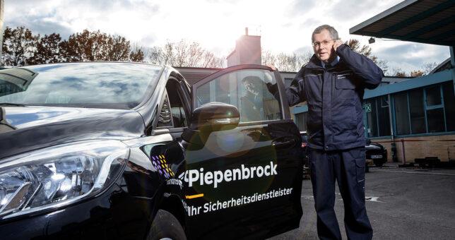 Piepenbrock Mitarbeiter mit dunkelblauer Hose und Jacke am telefonieren auf einem Parkplatz mit Piepenbrock-Auto und offener Tür
