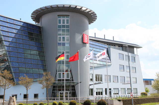 Moderne Unternehmenszentrale mit Glasfassade, rundem Turm und Flaggen vor dem Eingang bei sonnigem Wetter