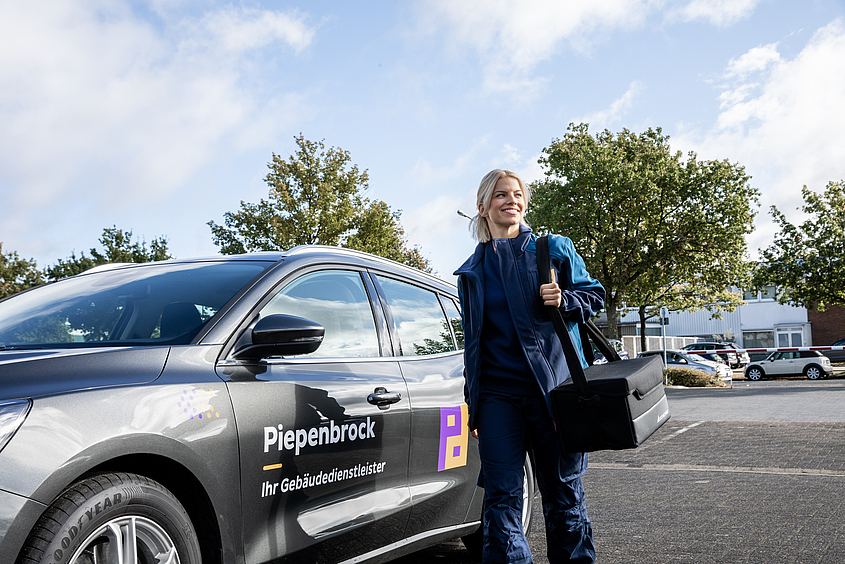 Frau in Arbeitskleidung und mit Tasche vor einem Piepenbrock-PKW