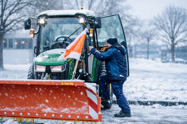 Piepenbrock Mitarbeiter steht neben Winterdienstfahrzeug mit Kehrbürste bei starkem Schneefall im Einsatz.